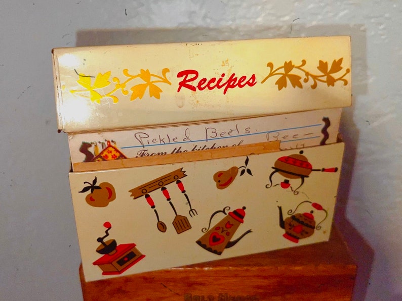 May include: Vintage recipe box with a cream-colored exterior and decorative kitchen-themed illustrations. The box is open, revealing recipe cards. The word "Recipes" is written in red on the lid. The box is made of metal.