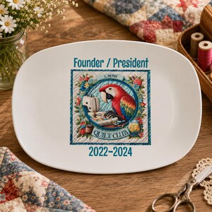 May include: A white oval ceramic plate featuring a central graphic of a colorful parrot sitting at a sewing machine. The text reads Founder / President and 2022-2024, with L MHH QUILT CLUB printed within a circular blue border surrounded by floral motifs.