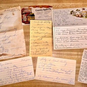 May include: A collection of handwritten recipes on various paper types, including index cards and aged paper. The recipes are for cakes, pies, and other baked goods. A vintage Country Apple Pie box is also visible.