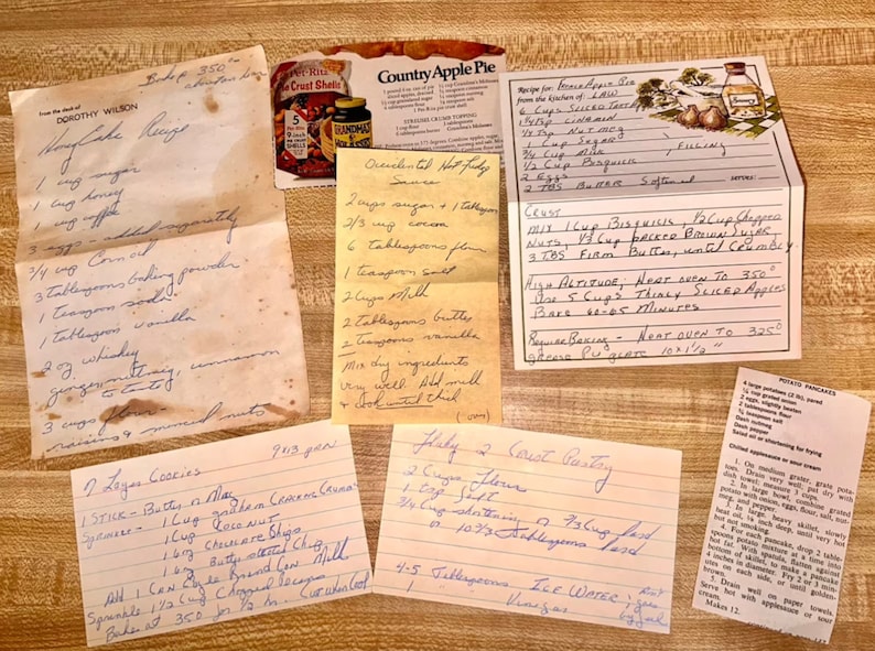 May include: A collection of handwritten recipes on various paper types, including index cards and aged paper. The recipes are for cakes, pies, and other baked goods. A vintage Country Apple Pie box is also visible.