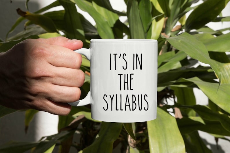 May include: White ceramic mug with black text that reads "It's in the syllabus".