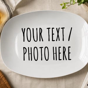 May include: A white oval ceramic serving platter featuring the black printed text "YOUR TEXT / PHOTO HERE" centered on the surface. The platter has a smooth, glossy finish and rounded edges, providing a clean and neutral base for custom designs.
