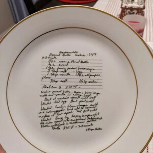 May include: A white ceramic plate with a handwritten peanut butter cookie recipe. The recipe includes ingredients like creamy peanut butter, brown sugar, all-purpose flour, salt, and baking soda. The instructions include beating the ingredients together, adding an egg, and baking the cookies at 375 degrees Fahrenheit for 7-8 minutes.
