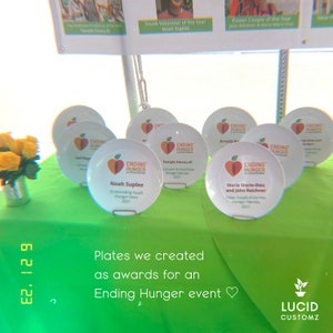 May include: A collection of white ceramic plates with a red apple logo and the text "Ending Hunger" on each plate. The plates are arranged on a green tablecloth with the text "Plates we created as awards for an Ending Hunger event" below them.