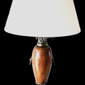 Puede incluir: Lámpara de mesa vintage con pantalla plisada blanca. La lámpara presenta un cuerpo de madera en forma de urna con detalles de latón y un remate decorativo. La base de la lámpara es una combinación de madera y latón.