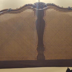 May include: A large wooden headboard featuring a curved top frame with carved detailing and three inset panels of woven cane webbing. The wood has a polished brown finish, and the cane panels are a natural light tan colour, creating a structured design.