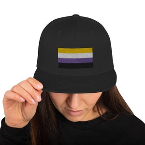 May include: A black baseball cap with an embroidered non-binary pride flag. The flag has yellow, white, purple, and black stripes.
