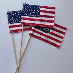 May include: Four miniature American flags with wooden sticks. The flags feature a blue square with white stars in the upper left corner and red and white stripes. The flags are arranged on a white surface.