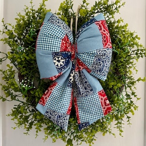 May include: A large decorative bow made of fabric with a patchwork pattern in shades of red, white, and blue, featuring gingham, paisley, and floral prints, attached to a circular wreath of green artificial leaves and small white flowers.