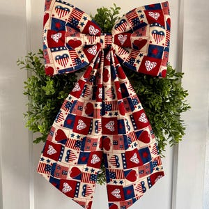 May include: A patriotic bow with a red, white, and blue pattern of hearts, stars, and flags. The bow is attached to a green wreath, creating a festive decoration for the home.
