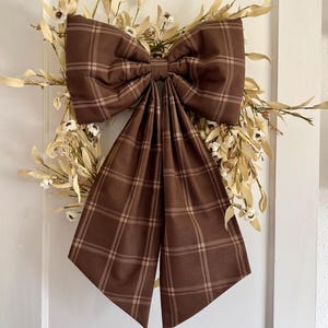 May include: A large brown fabric bow with a tan plaid pattern centered over a wreath made of dried cream-colored leaves and small white flowers. The bow features two wide loops and two long, hanging tails that drape downward over the foliage.