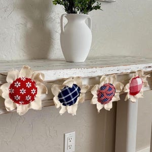 May include: A decorative garland featuring fabric flower shapes with various patriotic patterns. The garland includes red, white, and blue designs, including stars and stripes, and is displayed on a distressed white wooden table.