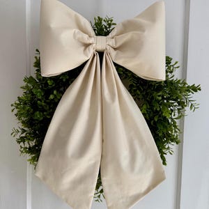 May include: A cream-colored fabric bow tied around a green boxwood wreath.