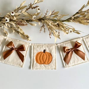 May include: A cream-colored banner with five square panels. Each panel features an embroidered orange pumpkin or a brown satin bow. The banner is decorated with dried floral stems, creating a festive autumn theme.