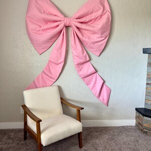 May include: A large, pink bow is mounted on a light gray wall. The bow has a voluminous design with long, flowing tails. Below the bow is a cream-colored armchair with wooden arms and legs, set on a brown carpet.