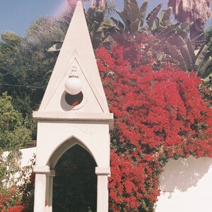 May include: A white, arched structure with a round light fixture on top, surrounded by vibrant red flowers and lush green foliage.