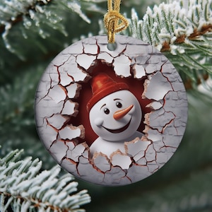 May include: A round Christmas ornament with a white cracked background. A smiling snowman with a red hat is peeking out of the crack.