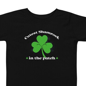 Puede incluir: Camiseta negra con un trébol verde con una cara sonriente. El texto "Cutest Shamrock * in the patch *" está impreso en la camiseta.