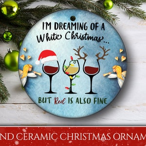 May include: Round ceramic Christmas ornament with a blue background and white text that reads "I'm dreaming of a white Christmas... but red is also fine". The ornament features three wine glasses, one with a Santa hat, one with reindeer antlers, and one with Christmas lights. Two angels are on either side of the glasses, each holding a trumpet.