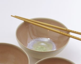 Könnte beinhalten: Drei Keramikschalen in verschiedenen Größen, alle mit einer hellrosa Glasur und einem weißen und grünen Design in der Mitte. Die Schalen sind auf einer weißen Oberfläche mit einem Paar Holzstäbchen angeordnet.