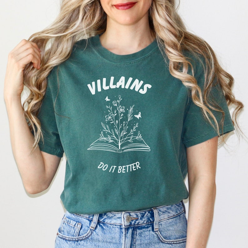Pode incluir: Uma camiseta verde-azulada com um gr&aacute;fico branco de um livro aberto com flores crescendo dele. O texto "VILLAINS DO IT BETTER" est&aacute; impresso na camiseta.