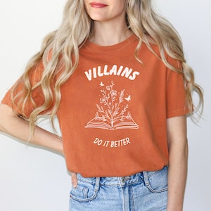Pode incluir: Uma camiseta cor ferrugem com um gr&aacute;fico branco de um livro aberto com flores e borboletas crescendo dele. O texto "VILLAINS DO IT BETTER" est&aacute; impresso na camiseta.