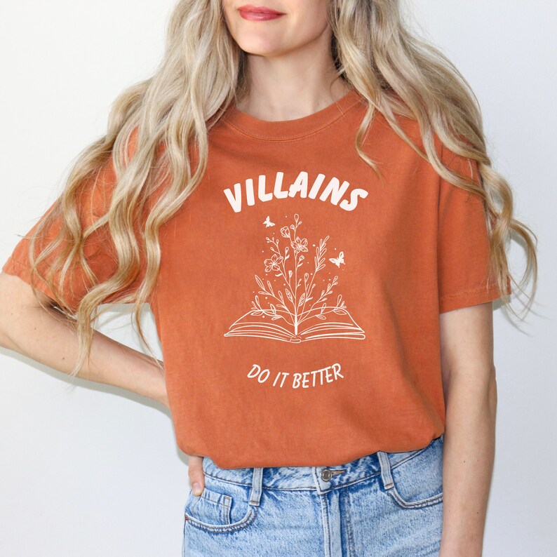 Pode incluir: Uma camiseta cor ferrugem com um gr&aacute;fico branco de um livro aberto com flores e borboletas crescendo dele. O texto "VILLAINS DO IT BETTER" est&aacute; impresso na camiseta.