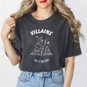 Pode incluir: Uma camiseta cinza escura com um design gr&aacute;fico branco de um livro aberto com uma flor crescendo dele. O texto "VILLAINS DO IT BETTER" est&aacute; impresso na camiseta.