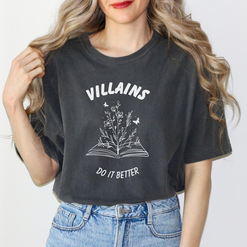 Pode incluir: Uma camiseta cinza escura com um design gr&aacute;fico branco de um livro aberto com uma flor crescendo dele. O texto "VILLAINS DO IT BETTER" est&aacute; impresso na camiseta.