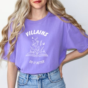 Pode incluir: Uma camiseta roxa com um design gr&aacute;fico branco de um livro aberto com flores e borboletas crescendo dele. O texto "VILLAINS DO IT BETTER" est&aacute; impresso na camiseta.
