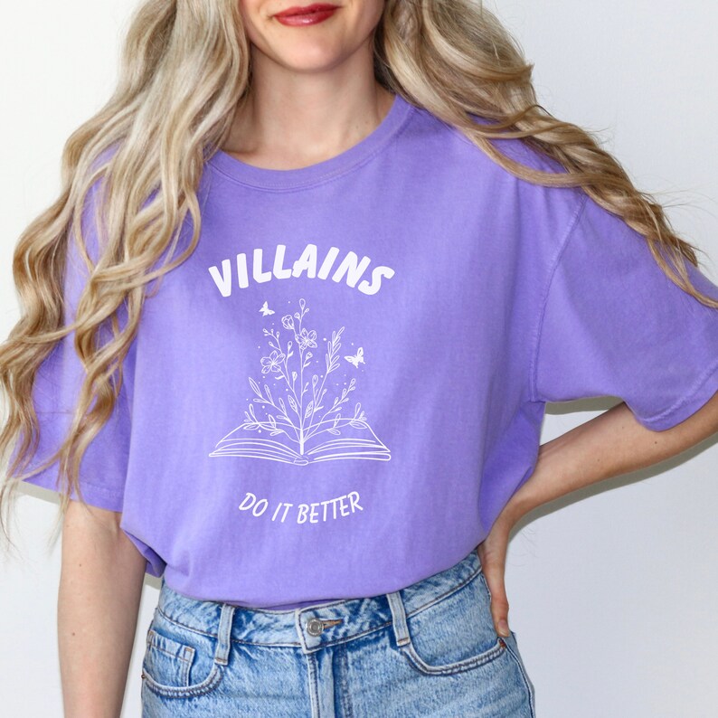Pode incluir: Uma camiseta roxa com um design gr&aacute;fico branco de um livro aberto com flores e borboletas crescendo dele. O texto "VILLAINS DO IT BETTER" est&aacute; impresso na camiseta.