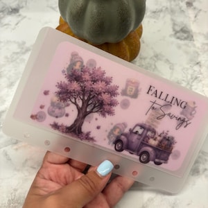 May include: A translucent rectangular plastic cash envelope with six punched holes along the left side. The design features a purple tree, a purple pickup truck carrying pumpkins, and the text FALLING to Savings in black script and serif font.
