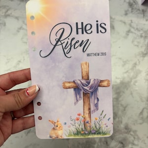May include: A planner insert with a watercolor design featuring a wooden cross draped with a purple cloth, a bunny, and the text "He is Risen" with the Bible verse Matthew 28:6. The background has a sunrise.