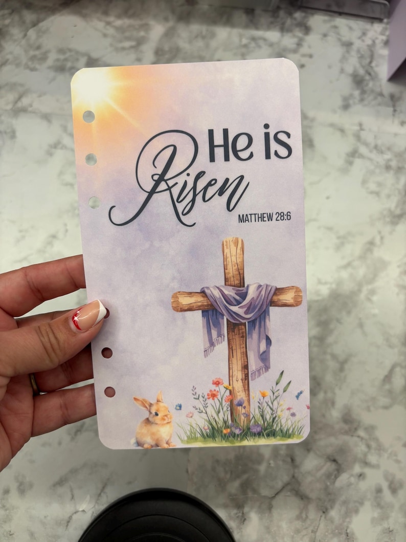 May include: A planner insert with a watercolor design featuring a wooden cross draped with a purple cloth, a bunny, and the text "He is Risen" with the Bible verse Matthew 28:6. The background has a sunrise.