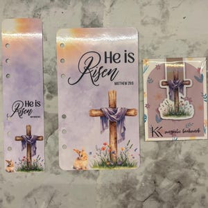 May include: Three Easter-themed paper products: a bookmark, a planner insert, and a magnetic bookmark. Each features a cross draped with a purple cloth, the words "He is Risen," and a bunny. The magnetic bookmark has the letter "K".