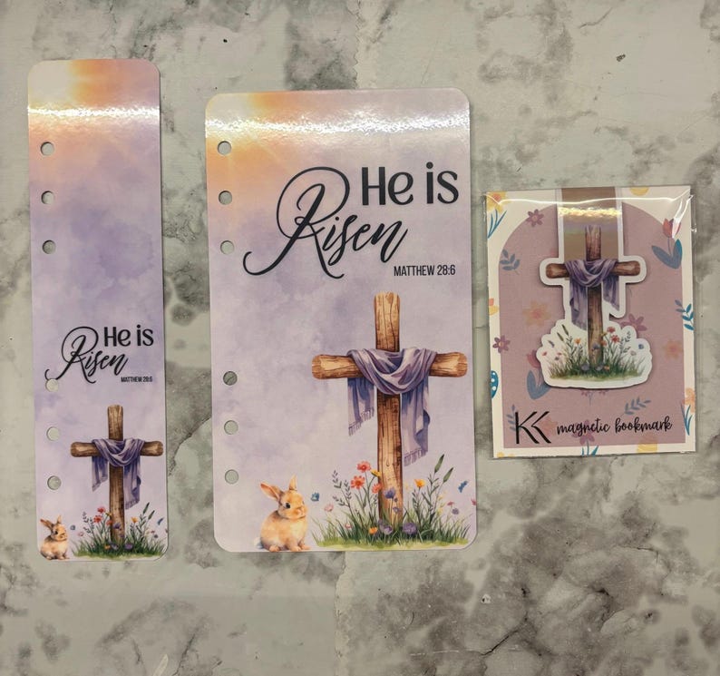 May include: Three Easter-themed paper products: a bookmark, a planner insert, and a magnetic bookmark. Each features a cross draped with a purple cloth, the words "He is Risen," and a bunny. The magnetic bookmark has the letter "K".