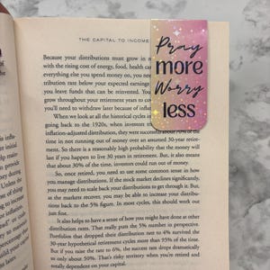 May include: A rectangular bookmark with a gradient of pink and gold, featuring the words "Pray more Worry less" in a handwritten style. The bookmark is placed in an open book.