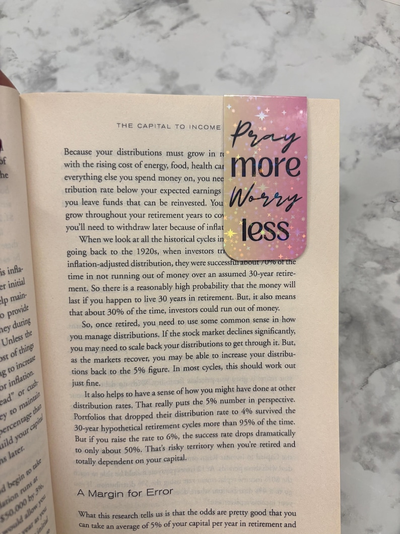 May include: A rectangular bookmark with a gradient of pink and gold, featuring the words "Pray more Worry less" in a handwritten style. The bookmark is placed in an open book.