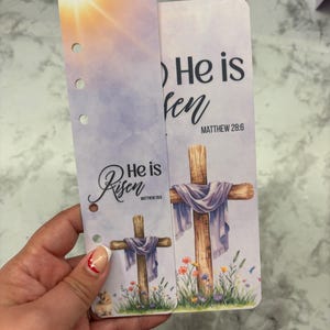 May include: Two-piece planner inserts with a religious theme. The inserts feature a cross draped with a purple cloth, surrounded by flowers, and the text "He is Risen" with Matthew 28:6. One insert has a sunrise background.