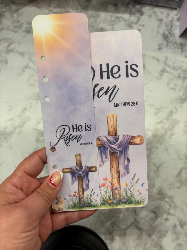 May include: Two-piece planner inserts with a religious theme. The inserts feature a cross draped with a purple cloth, surrounded by flowers, and the text "He is Risen" with Matthew 28:6. One insert has a sunrise background.