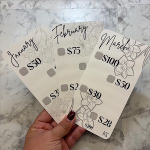 May include: Three white cards with floral designs and the months January, February, and March printed on them. Each card has a different amount listed, along with a series of gray squares. The cards are held in a hand.