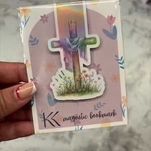 May include: A magnetic bookmark featuring a watercolor illustration of a wooden cross with a draped cloth and wildflowers. The bookmark is packaged in a clear plastic sleeve with a floral design and the text "magnetic bookmark".