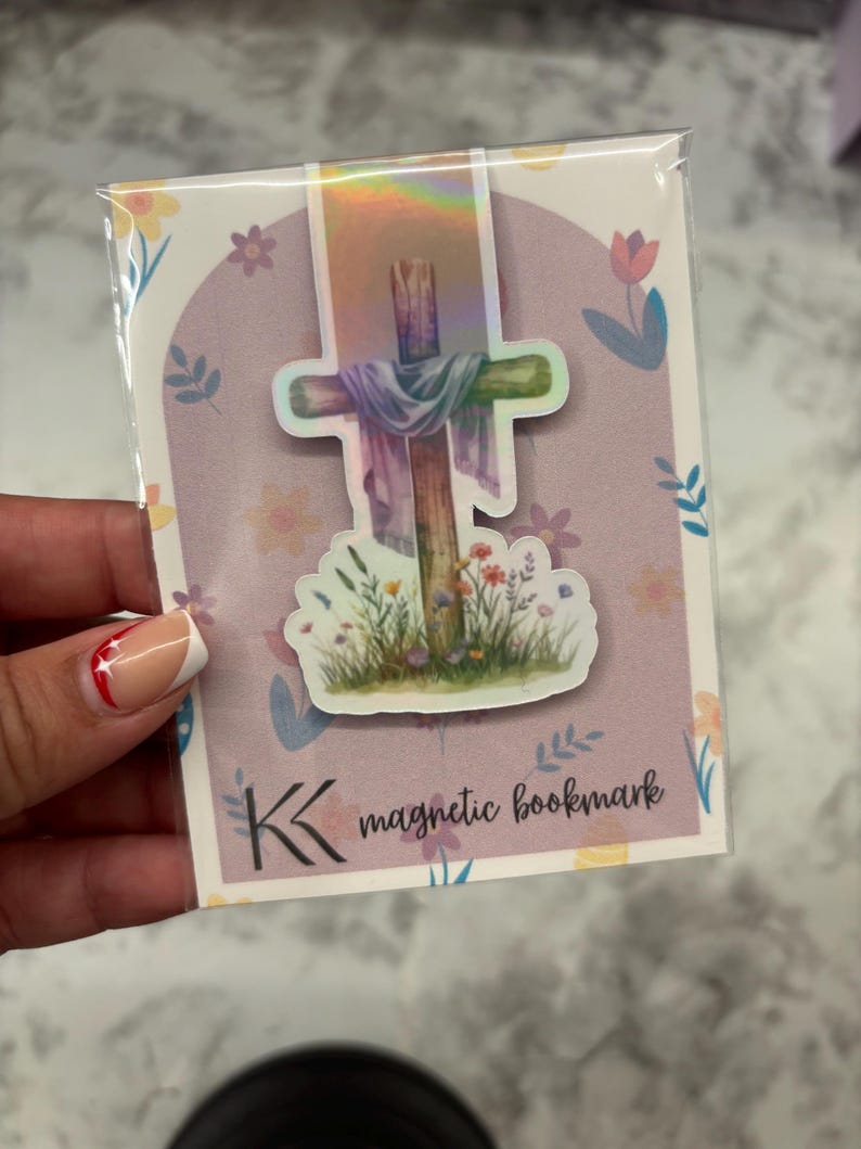 May include: A magnetic bookmark featuring a watercolor illustration of a wooden cross with a draped cloth and wildflowers. The bookmark is packaged in a clear plastic sleeve with a floral design and the text "magnetic bookmark".