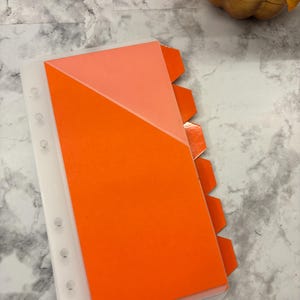 May include: A clear, plastic binder with a bright orange and peach-colored insert. The insert has a diagonal color block design. The binder has a series of tabs on the right side. The binder is on a marble-patterned surface.
