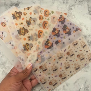 May include: A set of five translucent plastic binder inserts featuring various Halloween-themed patterns including cartoon ghosts, scarecrows, pumpkins, witches, and mummies. Each rectangular insert has a row of punched holes along the top edge.