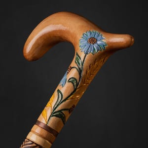 Pode incluir: Bengala de madeira entalhada à mão com um cabo curvo, apresentando desenhos florais e de folhas entalhados. A bengala é pintada com detalhes em azul, amarelo e verde. O eixo tem um padrão de folhas entalhadas e é de madeira clara.