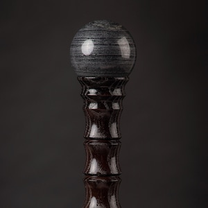 Op de afbeelding: Decoratieve wandelstok met een donkergrijze, bolvormige stenen kop. De schacht is gemaakt van donkerbruin hout met een glanzende afwerking en een gouden accent bij de basis. De stok heeft een uniek, elegant ontwerp.