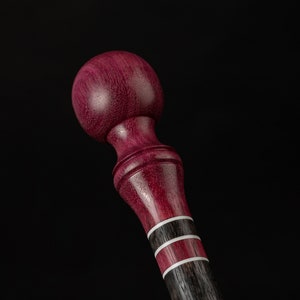 May include: A handcrafted wooden walking cane with a deep purple, rounded top and a dark gray shaft. The shaft features two white and red accent bands. The cane is set against a black background.