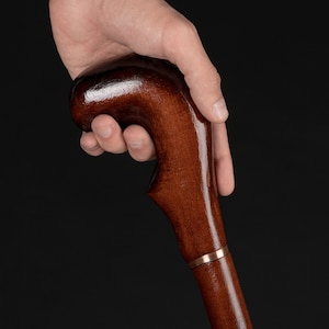 May include: A hand holds the top of a wooden walking stick with a brown finish. The stick has a gold band around it.