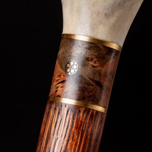 Puede incluir: Un bast&oacute;n de madera con un mango de hueso decorativo y una banda de lat&oacute;n. El bast&oacute;n tiene una veta de madera marr&oacute;n oscura y una incrustaci&oacute;n de lat&oacute;n con un dise&ntilde;o circular.